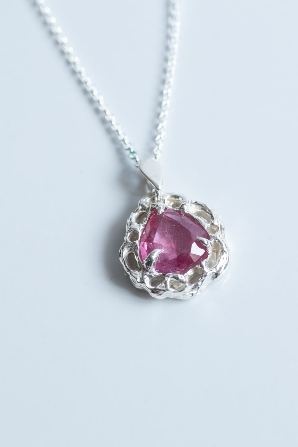 A silver pendant necklace featuring a heart-shaped pink gemstone surrounded by a textured, scalloped silver setting, suspended from a delicate silver chain on a light gray background.