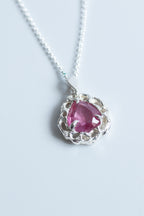 A silver pendant necklace featuring a heart-shaped pink gemstone surrounded by a textured, scalloped silver setting, suspended from a delicate silver chain on a light gray background.