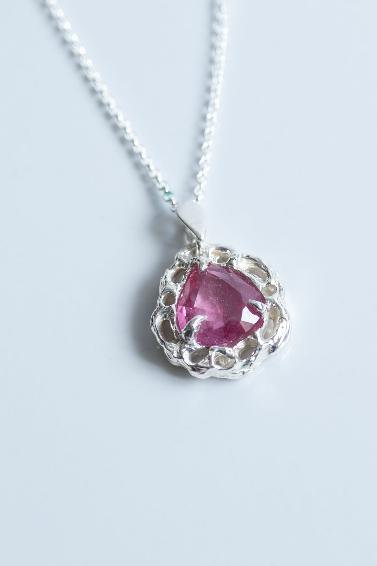 A silver pendant necklace featuring a heart-shaped pink gemstone surrounded by a textured, scalloped silver setting, suspended from a delicate silver chain on a light gray background.