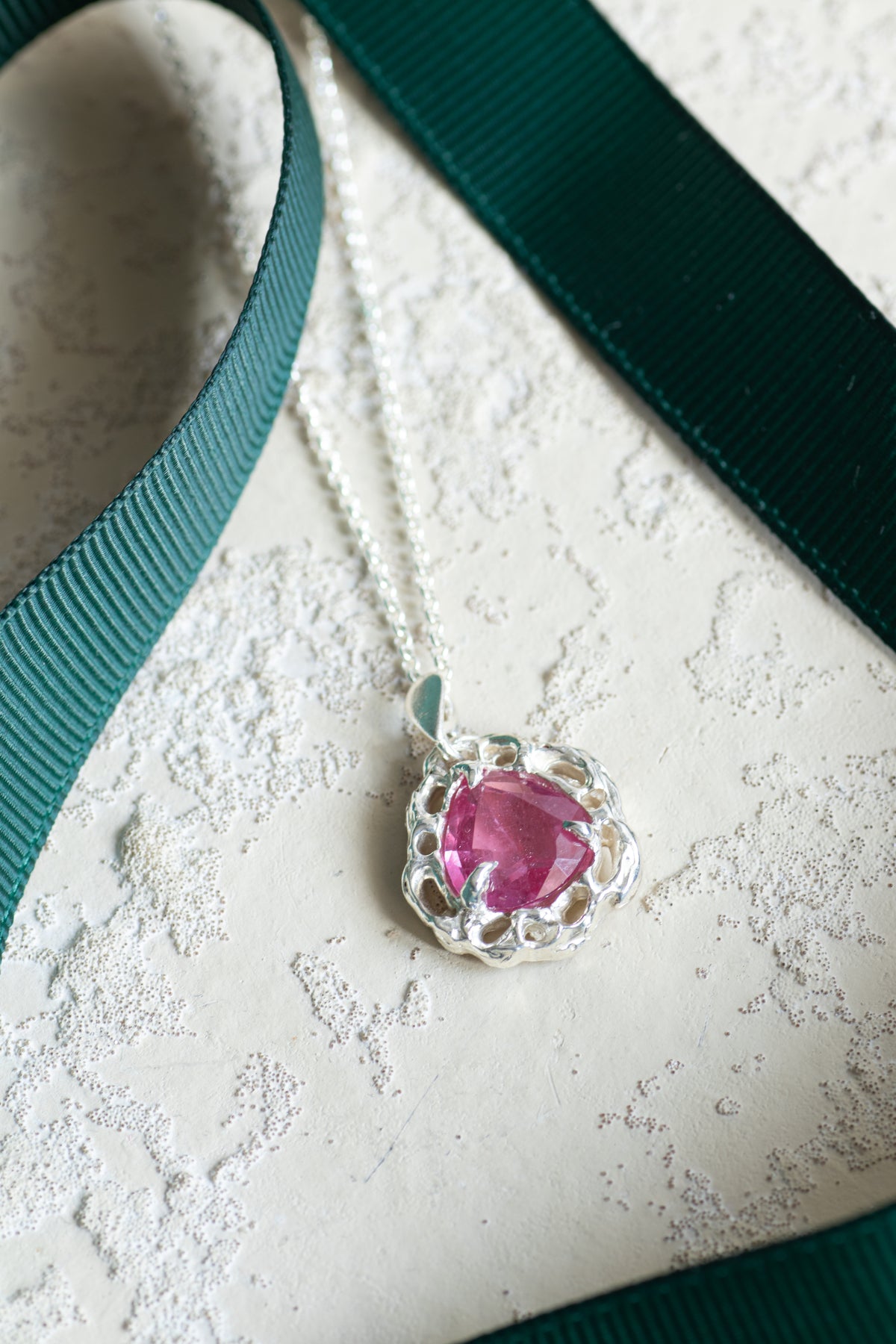 A silver pendant necklace with a heart-shaped pink gemstone, styled on a textured cream surface with a dark green ribbon.